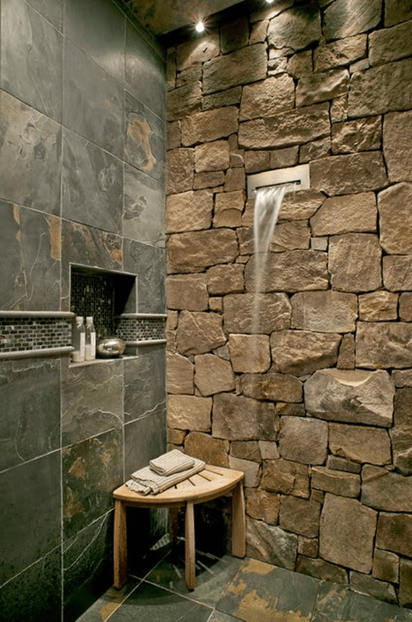 La imagen muestra un baño moderno y lujoso que combina elementos de piedra natural con paredes de azulejos elegantes. Destaca una ducha estilo cascada, iluminación ambiental y accesorios de baño elegantes dispuestos ordenadamente, ejemplificando la tranquilidad y el lujo. Si buscas este tipo de diseño para tu baño, en Construcciones Carmona podemos ayudarte a lograrlo, asegurando un acabado sofisticado y de alta calidad.