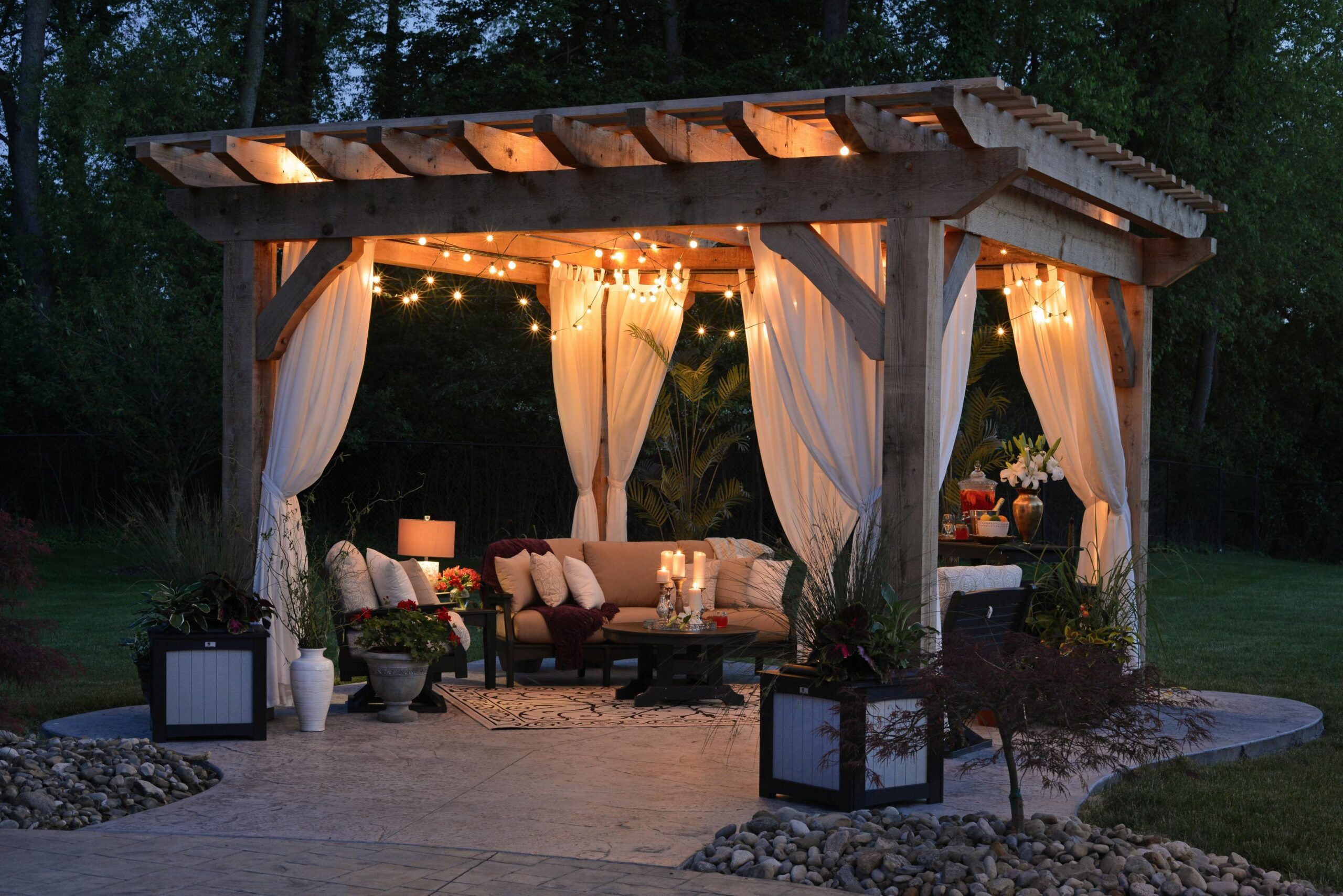 área de asientos al aire libre serena y bellamente iluminada durante la noche. Un cenador de madera con una estructura robusta y un techo de listones es la pieza central, adornado con elegantes cortinas blancas que se apartan suavemente. Luces de cuerda cálidas y brillantes se entrelazan alrededor de las vigas del cenador, proyectando una iluminación suave que realza el ambiente. Debajo del cenador hay un acogedor conjunto de asientos que consiste en un sofá mullido lleno de cómodos cojines, un espacio acogedor para la relajación o conversaciones íntimas. Una pequeña mesa frente al sofá sostiene objetos decorativos y velas, contribuyendo a la atmósfera cálida y acogedora. El suelo está cubierto con una alfombra intrincada que agrega textura y contraste al entorno. Rodeando este tranquilo espacio hay plantas y flores verdes exuberantes alojadas en elegantes maceteros, acercando los elementos de la naturaleza y agregando al atractivo estético. En el fondo, árboles altos bajo el cielo crepuscular contribuyen a una sensación de privacidad y reclusión.

Esta imagen captura un encantador área de asientos al aire libre bajo un cenador, iluminado por luces de cuerda; emana tranquilidad y calidez, invitando a los espectadores a un espacio donde la naturaleza se encuentra con la comodidad.