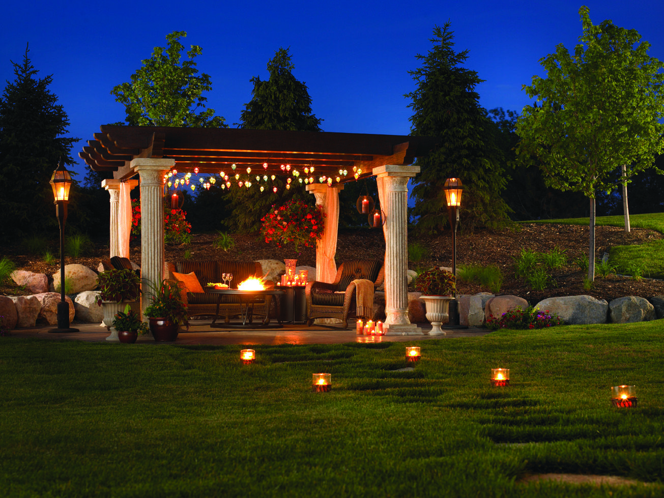 La imagen muestra un entorno al aire libre sereno y bellamente iluminado por la noche, con una pérgola, asientos cómodos, una fogata y vegetación circundante, evocando una sensación de relajación y tranquilidad.

Esta imagen captura una escena nocturna encantadora en un jardín donde varios elementos se combinan para crear una atmósfera de paz y relajación; es interesante debido a su armoniosa mezcla de iluminación natural y artificial que resalta la belleza del espacio al aire libre.
