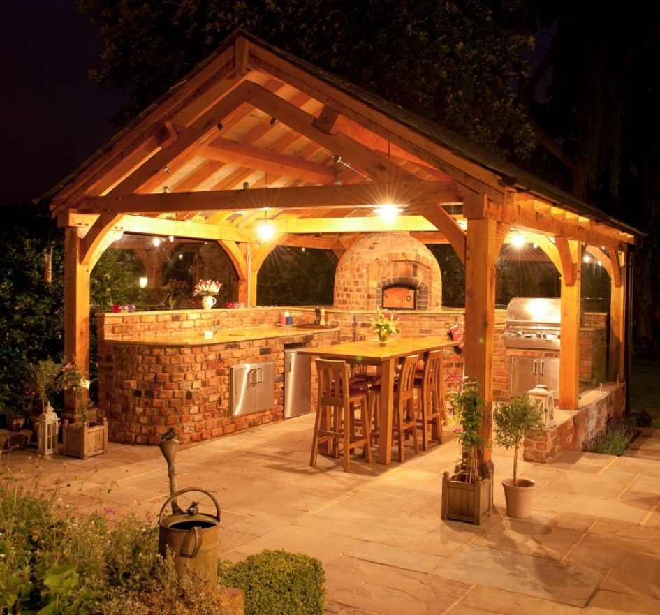 La imagen muestra una cocina al aire libre ubicada en un jardín durante la noche, iluminada por una luz cálida. La cocina está protegida bajo un cenador de madera con techo a dos aguas, sostenido por vigas robustas. Hay un mostrador de ladrillo a lo largo de un lado del cenador, equipado con un horno y una parrilla integrados. Cuatro taburetes de madera se encuentran frente al mostrador, invitando a una reunión acogedora. Plantas en macetas están colocadas alrededor del área, añadiendo al ambiente natural. El suelo está pavimentado con grandes piedras planas, complementando la estética rústica del entorno.

Esta imagen captura una cocina al aire libre acogedora, anidada en un jardín, resaltada por su iluminación cálida y diseño rústico que promete noches agradables al aire libre.
