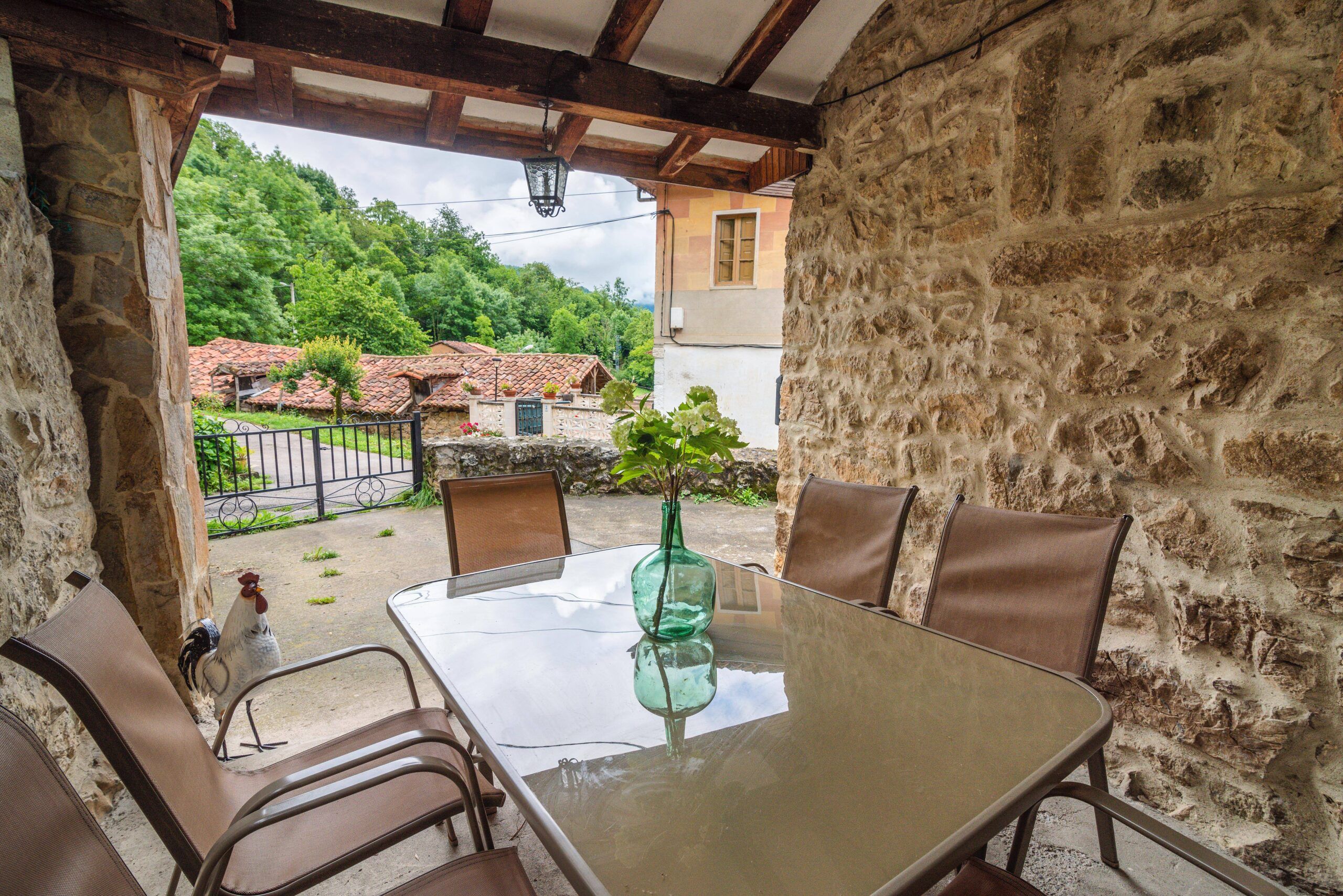 patio al aire libre serena y acogedora de una casa de piedra, mostrando una mezcla de elementos naturales y artificiales que crean una atmósfera armoniosa y relajante.

Para una descripción detallada adecuada para personas con discapacidad visual: El patio es un espacio tranquilo que invita a la relajación, con un suelo de baldosas que complementa la robustez de la arquitectura de piedra. La vegetación exuberante rodea el área, proporcionando un contraste refrescante con la solidez de la casa. Es un lugar ideal para disfrutar de la paz del entorno natural, sentir la brisa suave y escuchar los sonidos relajantes de la naturaleza. La combinación del encanto rústico de la arquitectura de piedra con la vegetación frondosa ejemplifica un espacio de vida al aire libre que invita a la calma y la contemplación.