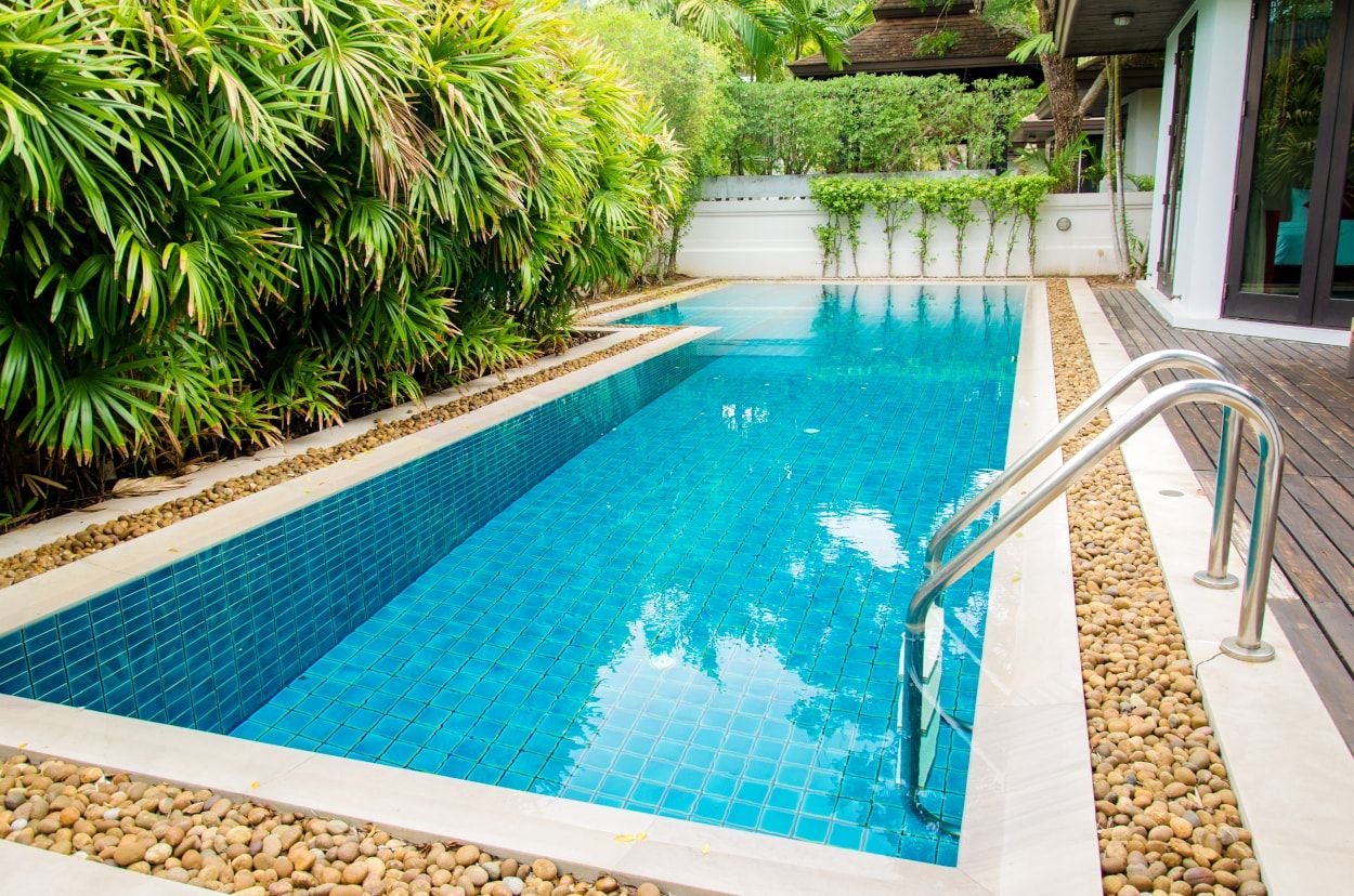 La imagen captura un área de piscina tranquila rodeada de vegetación exuberante y arquitectura moderna, ofreciendo una visión de un oasis privado que invita a la relajación y al disfrute. Si estás buscando crear un espacio sereno y acogedor al aire libre con una piscina cristalina, caminos de guijarros y arquitectura contemporánea, en Construcciones Carmona podemos ayudarte a hacer realidad ese sueño, asegurando que cada detalle se alinee con tus expectativas para el lugar perfecto de escape y tranquilidad en tu hogar.