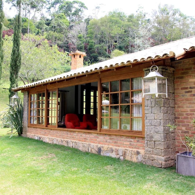  casa de un solo piso con paredes de ladrillo y ventanas grandes. Está rodeada por un jardín verde y tiene un fondo de árboles y vegetación. La casa tiene un aspecto acogedor y rústico, y las ventanas permiten ver sillas rojas en el interior. También hay una chimenea en el techo, lo que sugiere que podría haber una chimenea dentro. El cielo no es visible, pero la luz natural ilumina la escena, indicando que es de día.