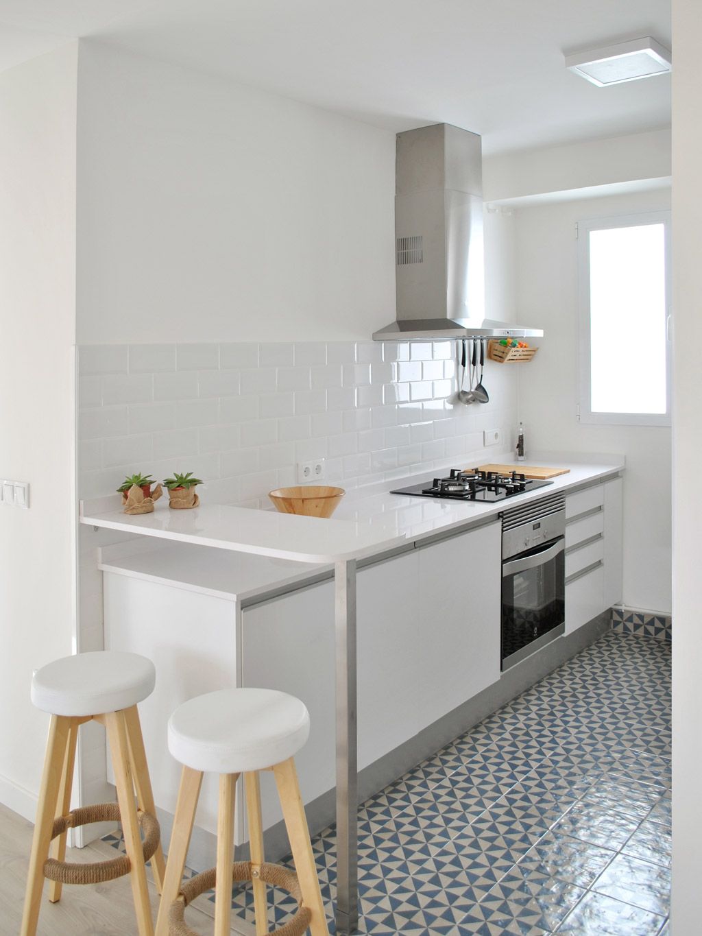 cocina moderna y luminosa con una estética minimalista. Hay una barra de desayuno con dos taburetes altos de madera y asiento blanco. Los gabinetes son blancos y lisos, con un diseño sin tiradores. La encimera también es blanca, contrastando con el protector contra salpicaduras de azulejos blancos. Hay una estufa y un horno integrados, con una campana extractora de acero inoxidable encima. El suelo está cubierto con baldosas en un patrón geométrico azul y blanco. Una ventana grande permite la entrada de luz natural, iluminando el espacio.