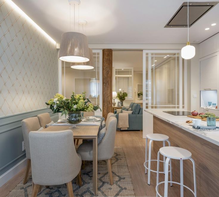 comedor y una cocina integrados con un diseño elegante y moderno. En el comedor hay una mesa de madera con sillas tapizadas en beige, decorada con flores en el centro. La pared tiene un papel tapiz de diseño geométrico sutil. Las lámparas colgantes sobre la mesa aportan una iluminación suave. La cocina tiene una isla con encimera blanca y taburetes blancos. Un separador con marcos blancos y un pilar de madera divide el comedor de la sala de estar, que se ve al fondo con sofás azul claro y decoraciones similares. El suelo es de madera clara y hay una alfombra bajo la mesa del comedor.