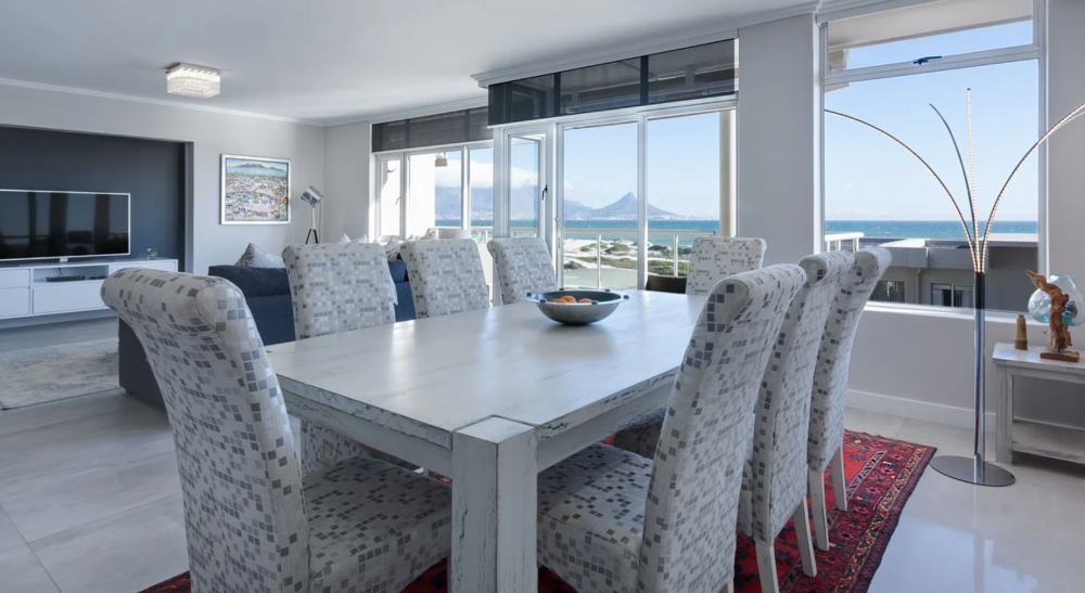 comedor y sala de estar con vista al mar. El comedor tiene una mesa de madera blanca y ocho sillas tapizadas con un patrón gris. Hay una alfombra roja debajo de la mesa. Grandes ventanales permiten la entrada de luz natural y ofrecen una vista panorámica del océano. La sala de estar cuenta con un sofá azul, una gran televisión y decoración moderna. La habitación tiene un diseño luminoso y espacioso, con paredes claras y pisos de baldosas.