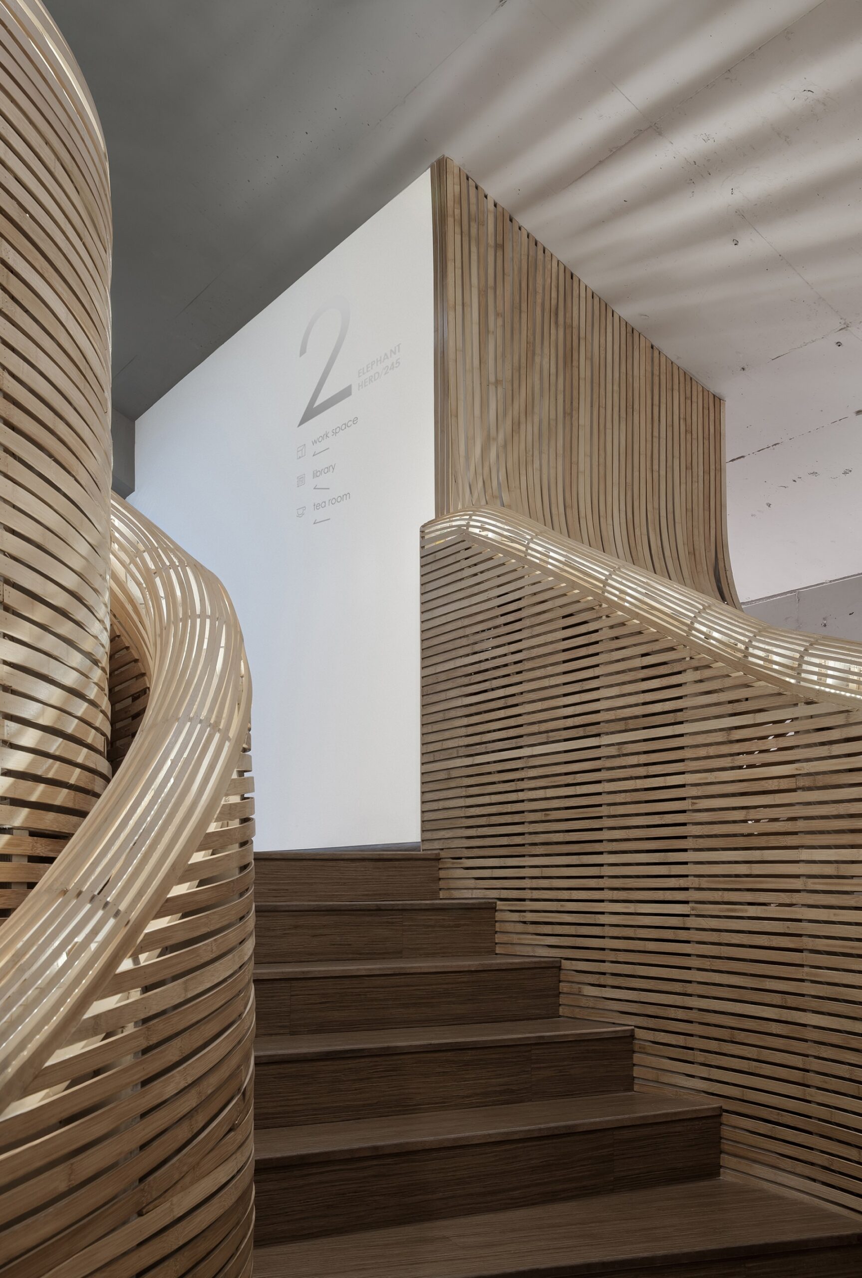 escalera moderna con un diseño curvo y detallado, hecha de madera con listones horizontales. Las paredes y el pasamanos están revestidos con estos listones, creando un efecto visual ondulado. En la pared blanca al fondo, se indican las direcciones a diferentes áreas: espacio de trabajo, biblioteca y sala de té. La iluminación es suave, destacando los contornos de la madera. El ambiente es contemporáneo y elegante.