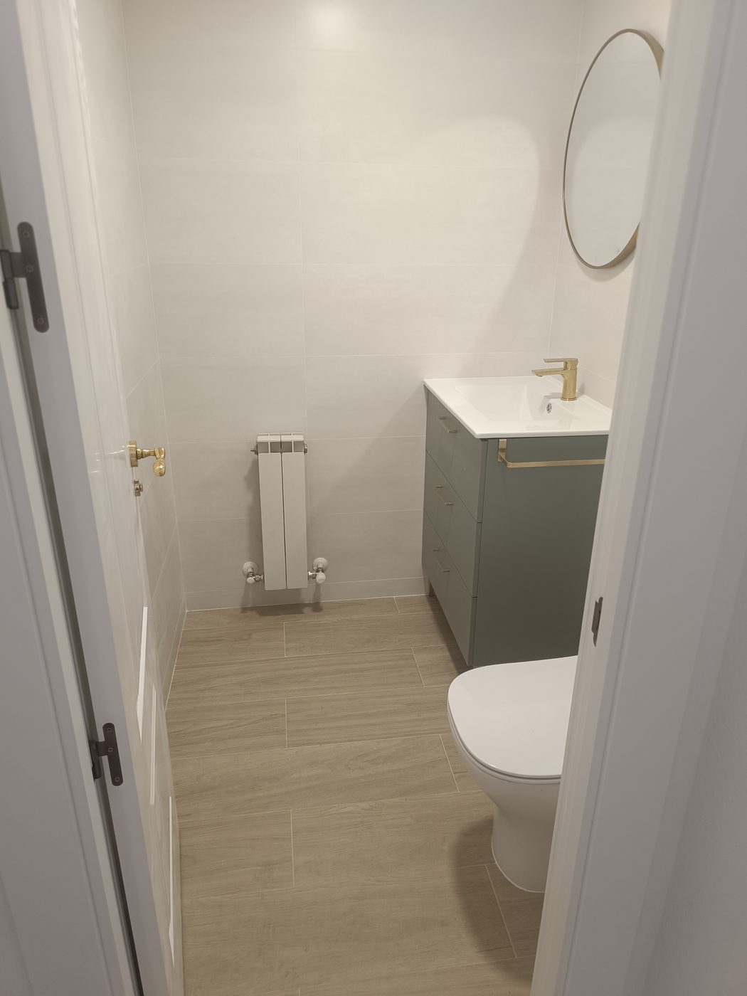 Aseo terminado en Moralzarzal con mueble verde salvia, grifería dorada, espejo redondo y suelo porcelánico imitación madera